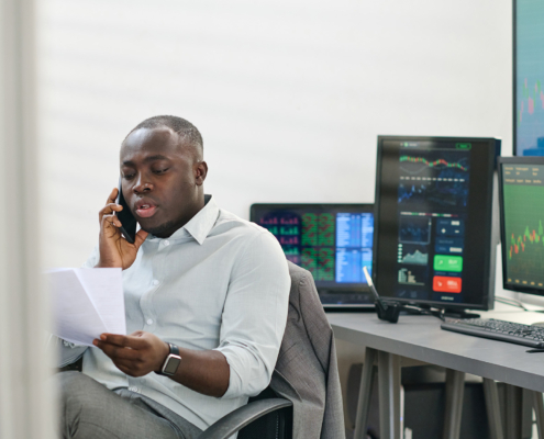 Young African American stock trading specialist wearing white shirt holding papers talking on phone